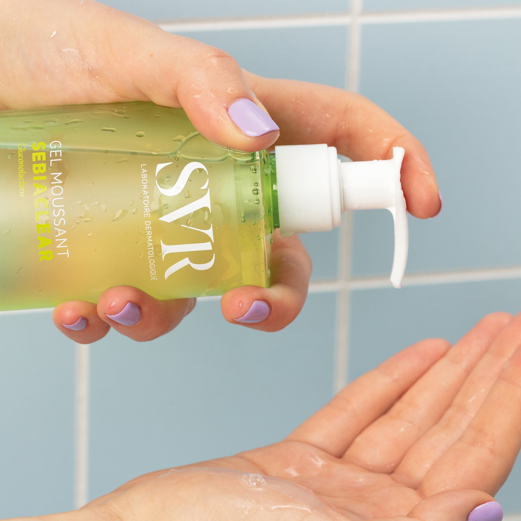 Hand with lavender nails dispensing SVR Sebiaclear Gel Moussant from a clear green pump bottle into an open palm.