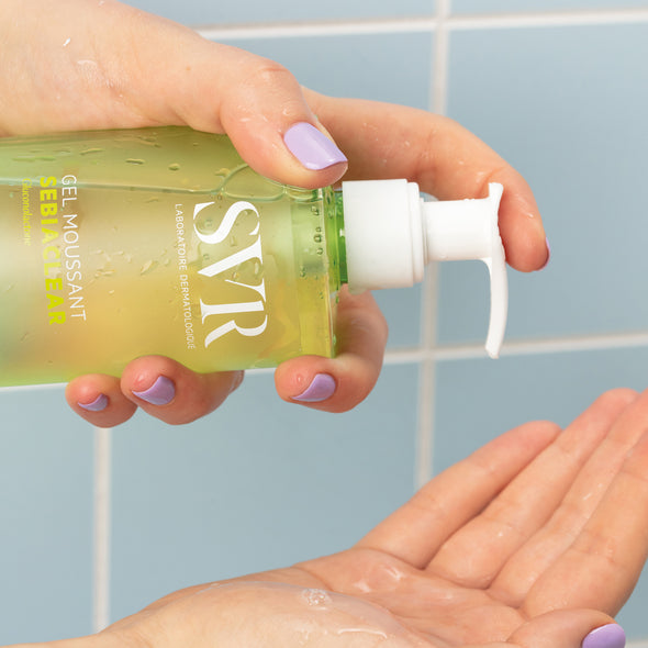 Hand with lavender nails dispensing SVR Sebiaclear Gel Moussant from a clear green pump bottle into an open palm.