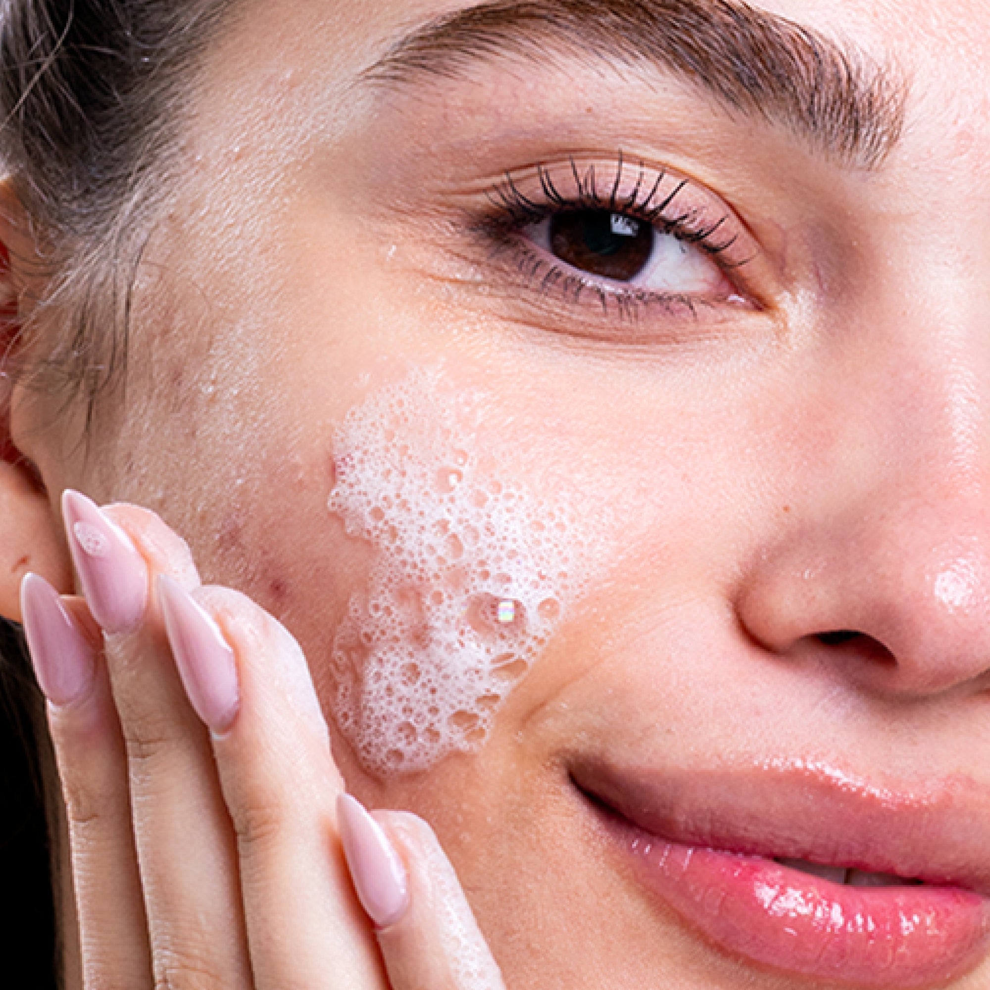 Close-up of a woman applying white foaming facial cleanser to her cheek with her hand.