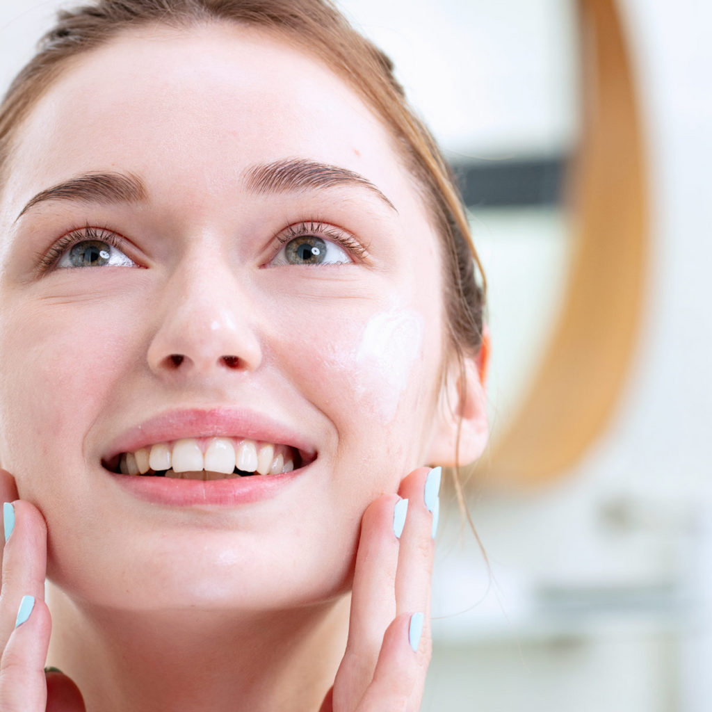 Young woman with clear skin applying white cream to her cheek with blue-painted nails.