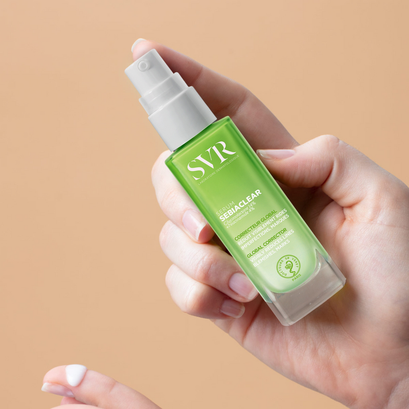 Hand holding SVR Sebiaclear Serum in green frosted glass bottle with white pump on beige background.