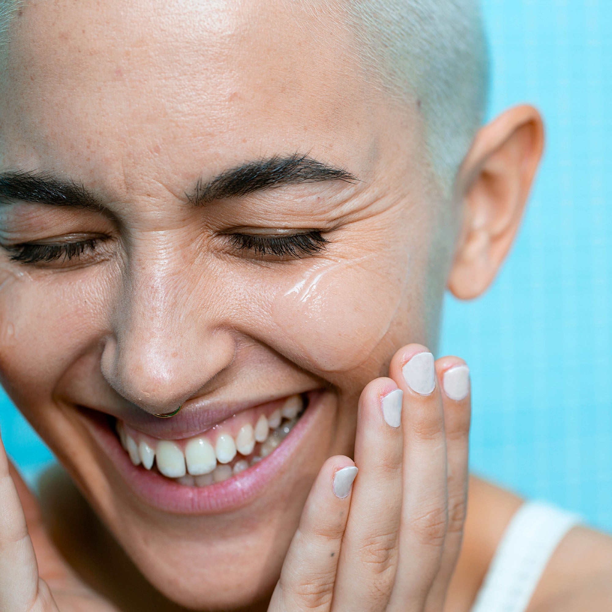 Smiling person with short blonde hair applying creamy cleansing balm to cheek against blue background.