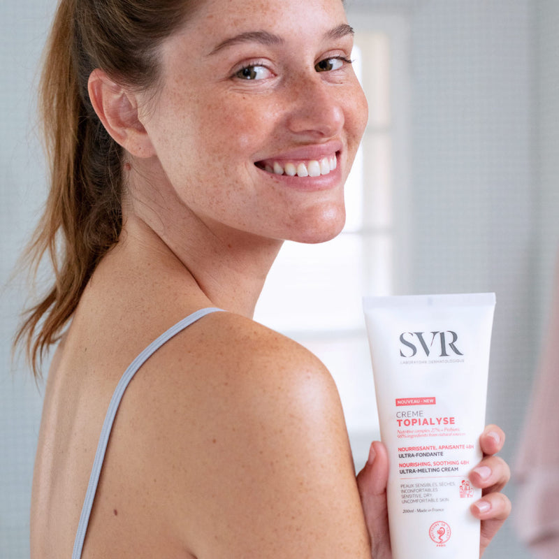 Woman smiling over shoulder holding white tube of SVR Topialyse cream with red and black text.